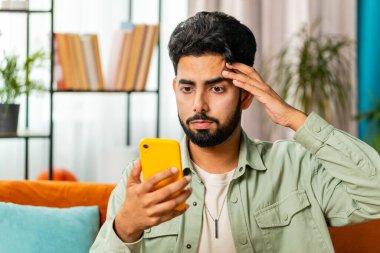 Hüzünlü Arap adam akıllı telefon daktilo kullanıyor, beklenmedik piyango sonuçlarını kaybediyor, kötü haber, servet kaybı, başarısızlık, teslim tarihi, virüs. Genç Hintli adam kanepede oturuyor.