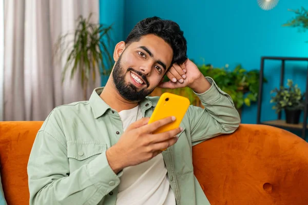 Indian man sits on couch uses mobile phone smiles at home living room apartment. Young Arabian guy texting share messages content on smartphone social media applications online, watching relax movie