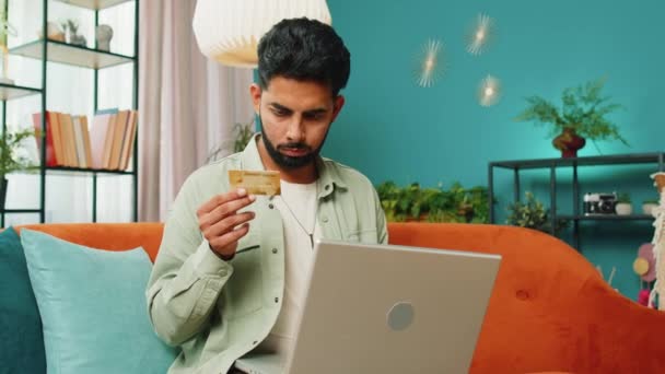 インドの男性は クレジットバンクのプラスチックカードとラップトップコンピュータを使用して お金を転送しながら オンラインショッピングを購入し 現代のホームルームアパートでインターネット上の食品配達を注文します ソファーに座っている男 — ストック動画