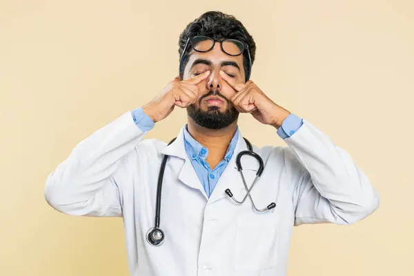 Exhausted tired Indian young doctor cardiologist man takes off glasses, feels eyes pain, being overwork burnout from long hours working. Sleepy exhausted guy rubbing eyes isolated on beige background