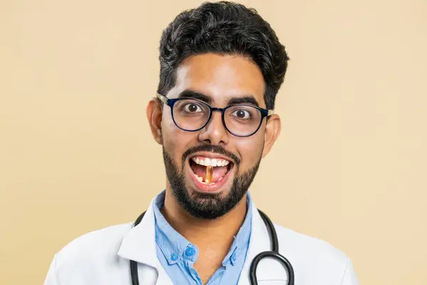 Smiling Indian doctor man puts of tablet pill vitamin D or omega-3 yellow capsule into mouth, recommends drugs immunization cure treatment. Health care. Apothecary pharmacist on beige background