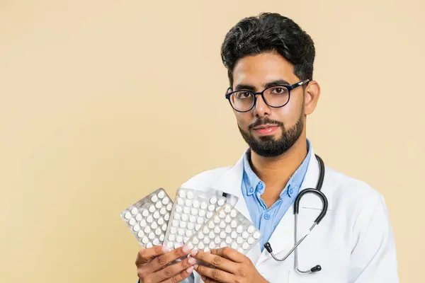 Indian doctor cardiologist man shows lot of tablet pills in blisters, recommends drugs vitamin antibiotic cure treatment. Health care. Arabian apothecary pharmacist guy isolated on beige background
