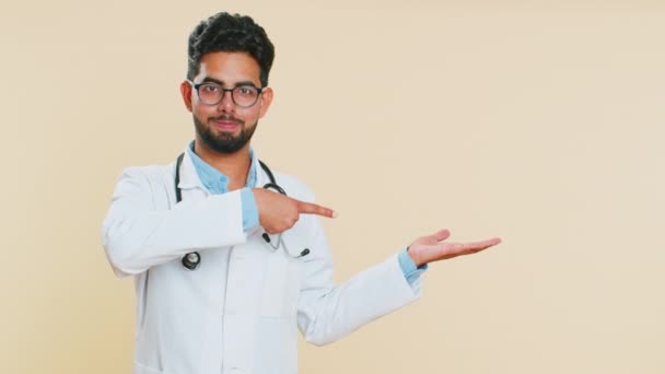インドの若い医師の心臓病学者男性は親指を立てて 商業割引 コピースペースのための広告領域を指差します ベージュの背景に隔離されたアラビアの薬局の男 — ストック動画