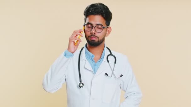 無関心なインドの医師心臓専門医は スマートフォンで退屈な話をすることに迷惑をかけています 疲れた睡眠アラビアの薬局の男フリーランサーベージュの背景に友人と携帯電話で話す — ストック動画