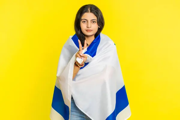 Indian smiling young woman holding Israel national flag, showing victory sign against war, hoping for success and win, doing peace gesture. Independence, freedom, national holiday Arabian Hindu girl