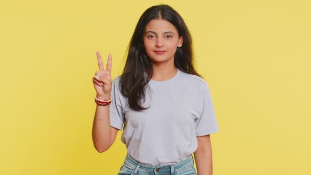 快乐的印度年轻女子展示出胜利的迹象 希望成功和胜利 做一个和平的姿态 带着友善乐观的微笑 庆祝胜利 在工作室黄色背景上被隔离的阿拉伯女孩 — 图库视频影像