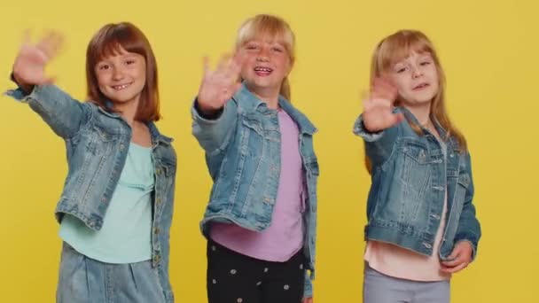 10代の女の子はカメラで優しい笑みを浮かべて手を振って挨拶や別れをジェスチャーで手を振って もてなしの表情で歓迎します 妹たちよ スタジオで3人の兄弟子供黄色の背景 — ストック動画