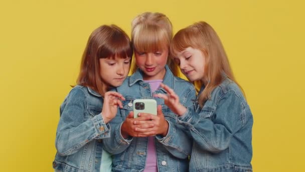 十代の女の子は 携帯電話の入力のブラウジングを使用すると言うすごいはい偉大な勝利良いニュースのお祝いを発見し オンラインゲームをプレイ 妹たちよ 黄色の背景をした3人の兄弟 — ストック動画