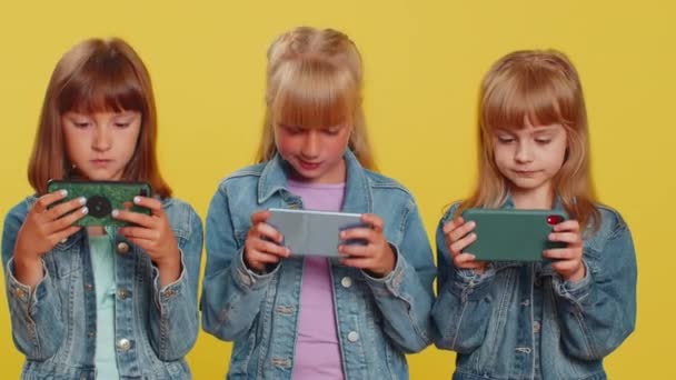 忧心忡忡的少女们兴致勃勃地在智能手机上玩赛车或射击在线视频游戏 孩子们的妹妹们使用智能手机小应用程序与驱动器模拟器 三个兄弟姐妹的孩子 — 图库视频影像