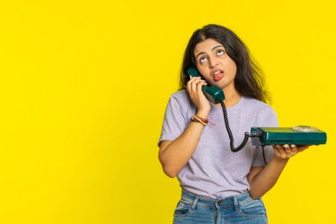 80 'lerin telli vintage telefonundan konuşan, aptal suratlar yapan sıkıcı hikayelerden bıkmış, iletişim konuşmalarıyla ilgilenmeyen, sıkılmış Hintli bir kadın. Sarı arka planda Arap bir kız