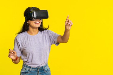 Kulaklık uygulaması kullanarak simülasyon oyunu oynayan heyecanlı Hintli genç kadın. Sanal gerçeklik 3D 360 videosu izliyoruz. VR gözlüklü Arap kız sarı arka planda izole edilmiş. Gelecek teknolojisi