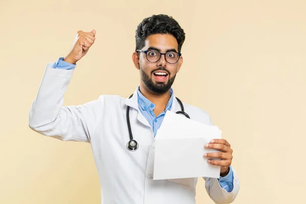 Indian young doctor cardiologist man open envelope take out letter reads it feel happy. Career growth advance promotion, long-awaited invitation great news. Arabian apothecary guy on beige background