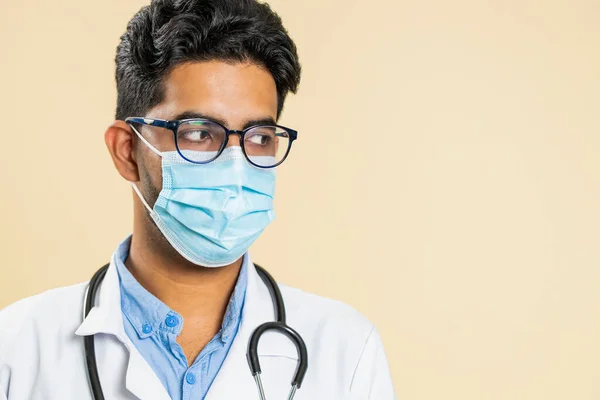 Face portrait of Indian young doctor cardiologist man lab worker in glasses looking at right empty place advertising promotion area. Arabian apothecary pharmacy guy in medical mask on beige background