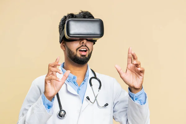 Excited happy Indian doctor cardiologist man using headset helmet app, watching virtual reality 3D 360 video. Arabian apothecary guy in VR goggles on beige background. Future medicine technology