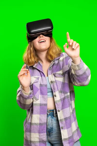 Excited happy woman using headset helmet app to play simulation game. Watching virtual reality 3D video. Redhead girl in VR goggles isolated on green chroma key background. Future technology. Vertical