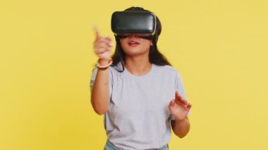 Kulaklık uygulaması kullanarak simülasyon oyunu oynayan heyecanlı Hintli genç kadın. Sanal gerçeklik 3D 360 videosu izliyoruz. VR gözlüklü Arap kız sarı arka planda izole edilmiş. Gelecek teknolojisi