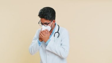 Koruyucu maskeli sağlıksız Hintli genç doktor mevsimsel grip belirtileriyle öksürüyor. Arap eczacı kendini hasta, alerji, ateş ya da bej arka planda izole edilmiş viral enfeksiyon hissediyor.