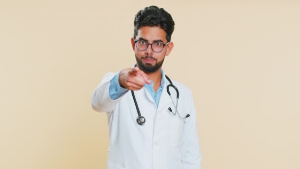 快乐而严格的印度年轻心脏病医生男人指着相机 向你表明 以米色背景孤立的阿拉伯药剂师微笑的家伙 — 图库视频影像