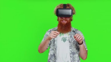 Heyecanlı sakallı adam, simülasyon oyunu oynamak için kulaklık uygulaması kullanıyor. Sanal gerçeklik 3D 360 videosu izliyoruz. VR gözlüklü kızıl saçlı adam yeşil krom anahtar arka planda izole edilmiş. Gelecek teknolojisi