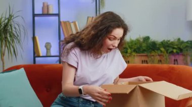 Evdeki kanepede oturan mutlu, genç bir kadın teslimat paketini açıyor. Gülümseyen tatmin olmuş kız alışverişci internet müşterisi karton kutu açıyor hızlı posta yoluyla satın alma hediyesi alıyor