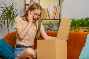 Evdeki kanepede oturan mutlu, genç bir kadın teslimat paketini açıyor. Gülümseyen tatmin olmuş kız alışverişci internet müşterisi karton kutu açıyor hızlı posta yoluyla satın alma hediyesi alıyor