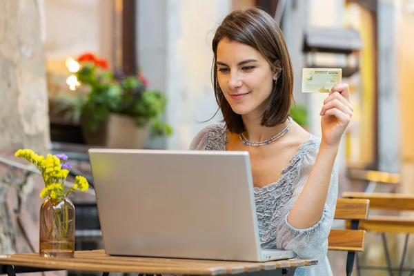 Young woman using credit bank card laptop computer while transferring money, purchases online shopping, order food delivery booking hotel room outdoors. Girl tourist sitting at table in city cafeteria