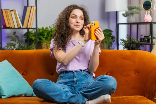 Portrait of woman sitting on sofa uses mobile phone smiling at home living room apartment. Young girl texting share messages content on smartphone social media applications online watching relax movie