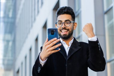 Mutlu Hintli işadamı cep telefonuyla kutlama yapıyor. İyi haber kazan, piyango zaferi kazan, online açık hava. Şehir merkezinde Arap Hindu serbest çalışanı. İş adamı yaşam tarzı