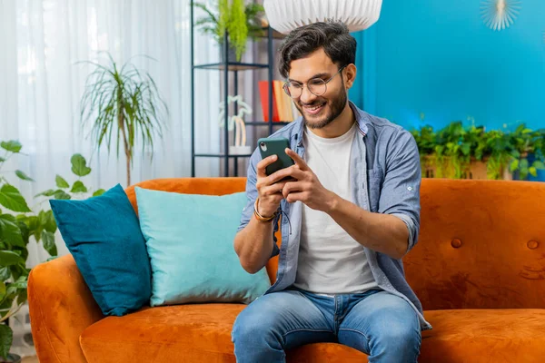Indian man sits on couch uses mobile phone smiles at home living room apartment. Young Arabian guy texting share messages content on smartphone social media applications online watching relax movie
