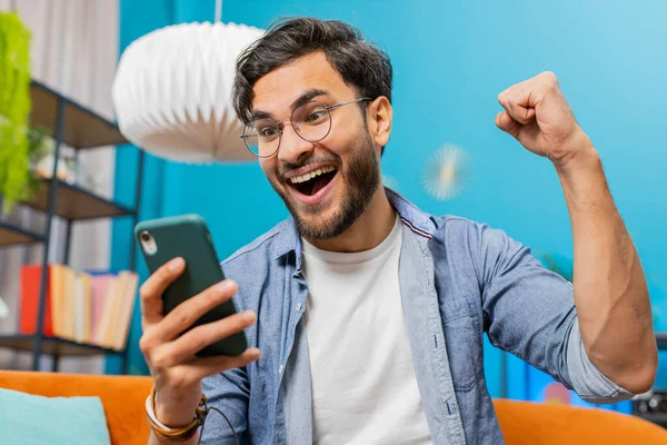Happy man using mobile smartphone typing browsing say Wow yes, celebrating success victory, winning lottery jackpot goal achievement play game good positive news, triumph. Guy at home in room on sofa