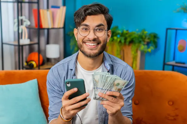Planning budget. Rich happy Indian man use smartphone calculator app count money cash, calculate domestic bills at home. Guy satisfied of income earnings win saves money for planned vacation gifts