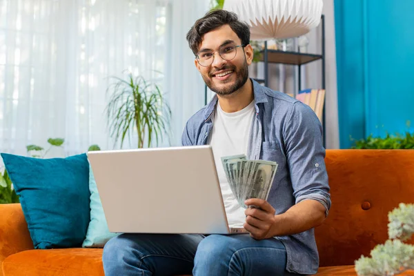 Rich happy Indian man counting money cash use laptop computer calculate domestic bills at home room. Planning budget. Arabian guy satisfied of income earnings, saves money for planned vacation, gifts