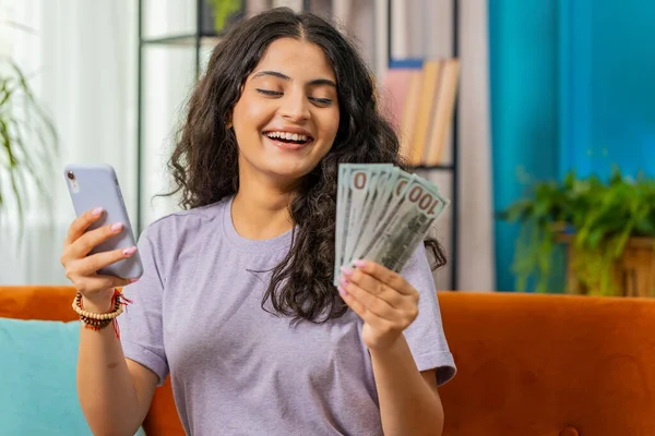 Planning budget. Rich happy smiling Indian woman use smartphone calculator app count money cash, calculate domestic bills at home. Girl satisfied of income earnings win saves money for vacation gifts