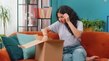 Kızgın, hoşnutsuz Hintli kadın paketlerini açıyor üzgün ve kafası karışmış bir şekilde çevrimiçi bir dükkandan gelen yanlış teslimat, kötü kalite bozuk ev alımı. Kız içerde kanepede yatıyor.