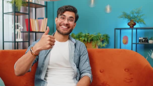 同じように ハッピーエキサイティングなインド人男性は 良いニュースの肯定的なフィードバックをサインのように親指を示すカメラで承認して見ています 若いアラビアの男は 自宅のリビングルームでソファーに座っています — ストック動画