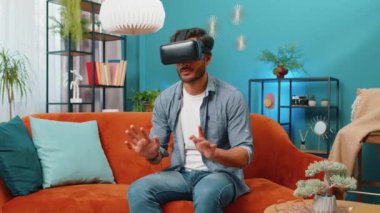 Hintli adam sanal gerçeklik teknolojisi VR uygulama başlığı kullanarak sanal klavyeye yazı yazarak, evdeki dairede 3D video oyunu simülasyonu yaparak. Kanepede oturan gözlüklü adam.