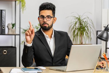 Indian young businessman working on laptop computer shakes finger and saying No be careful scolding and giving advice to avoid danger mistake disapproval sign at home office. Confident freelancer man