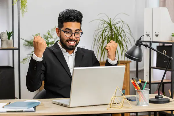 Happy Indian business man working on laptop at home office desk typing browsing celebrating success victory, winning lottery jackpot goal achievement play game good positive news, triumph