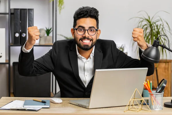 Happy amazed young Indian businessman working on laptop shocked by sudden victory, game winning, lottery goal achievement good career news. Excited manager man freelancer at home office workplace desk