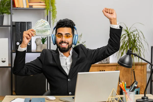 Indian businessman holding fan of cash money dollar banknotes celebrate, dance, success career, lottery jackpot game winner, big income, wealth at home office workplace desk. Remote distant working