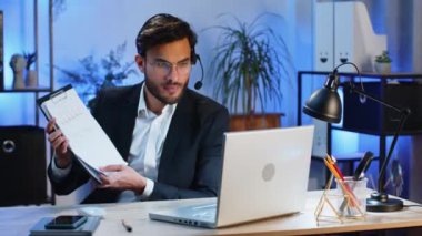 Kulaklık takan Hintli işadamı, serbest çalışan, çağrı merkezi destek operatörü yardım hattı, ofiste diyagram grafik istatistiklerini gösteren iş arkadaşı iletişim desteği ile konuşmak,