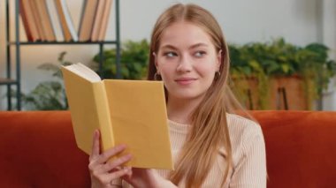 Genç sarışın kadın ilginç kitaplar okuyor edebiyatın tadını çıkarıyor rahat koltukta dinleniyor. Huzurlu, gülümseyen, neşeli kız evinde, oturma odasında, koltukta oturuyor.