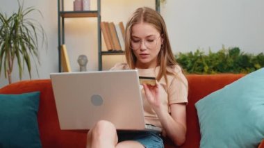 Genç sarışın kadın kredi kartı ve dizüstü bilgisayar kullanarak para aktarıyor, online alışveriş yapıyor, eve yemek siparişi veriyor. Oturma odasında kanepede oturan yetişkin kız.