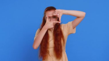 Genç yetişkin kadın fotoğrafçı elleriyle resim çerçevesi çiziyor, parmaklarından bakıyor ve ilginç bir ana odaklanıyor, zoom 'u taklit ediyor ve güzel görüntüler kesiyor. Mavi arka planda kızıl saçlı kız