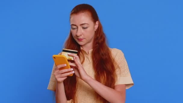 クレジットバンクカードとスマートフォンを使用して オンラインショッピング 支払いを購入するシンセレアの若い女性顧客 金融とインターネット 青い背景で孤立した赤毛の少女 — ストック動画