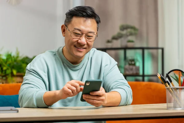Asian man sits on couch uses mobile phone smiles at home room apartment. Middle-aged Chinese guy texting share messages content on smartphone social media applications online watching relax movie