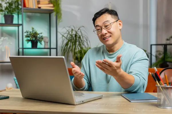 Asian man looking at laptop camera, making video webcam conference call with friends or family, enjoying pleasant conversation at home apartment. Guy laughing, waving hello sits on couch at table desk
