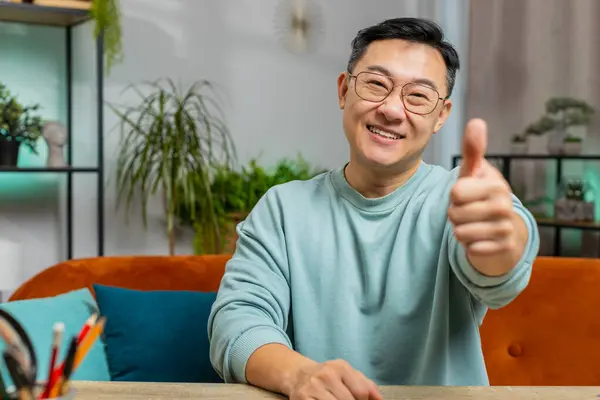 Like. Happy excited mature Asian man looking approvingly at camera showing thumbs up like sign positive something good great news positive feedback. Middle-aged Chinese guy sits on couch at home room