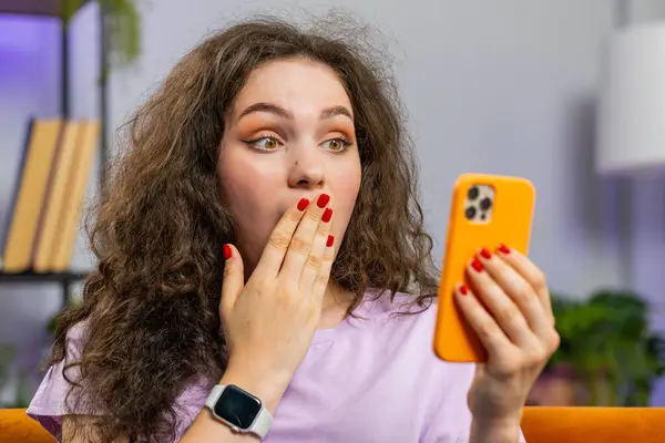 Oh my God Wow. Excited happy young woman use smartphone typing browsing celebrating success winning, lottery jackpot goal achievemen, good news, victory. Caucasian girl holds mobile phone at home room