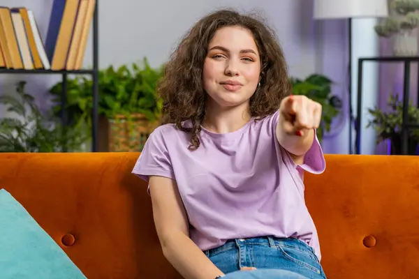 Hey you. Young woman smiling excitedly and pointing to camera, choosing lucky winner, indicating to awesome you, inviting, approve. Portrait of adult girl at modern home living room sitting on couch